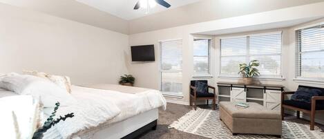 Une chambre à coucher comprenant un lit, un bureau, deux chaises, un ventilateur de plafond et un téléviseur fixé au mur.