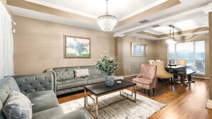 A living room with a sectional sofa, a coffee table, a dining table with chairs, a chandelier, and a large window with blinds. - 4BDR+PrivatePool+Game Room+BBQ+FirePit+Fits12 (Corpus Christi)
