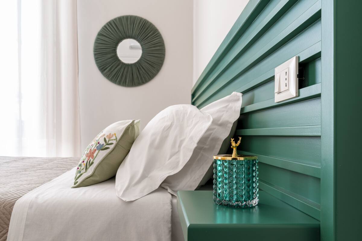 Un lit bien fait, avec du linge de lit blanc et des coussins décoratifs, une table de chevet verte agrémentée d’un objet décoratif, et un miroir fixé au mur.