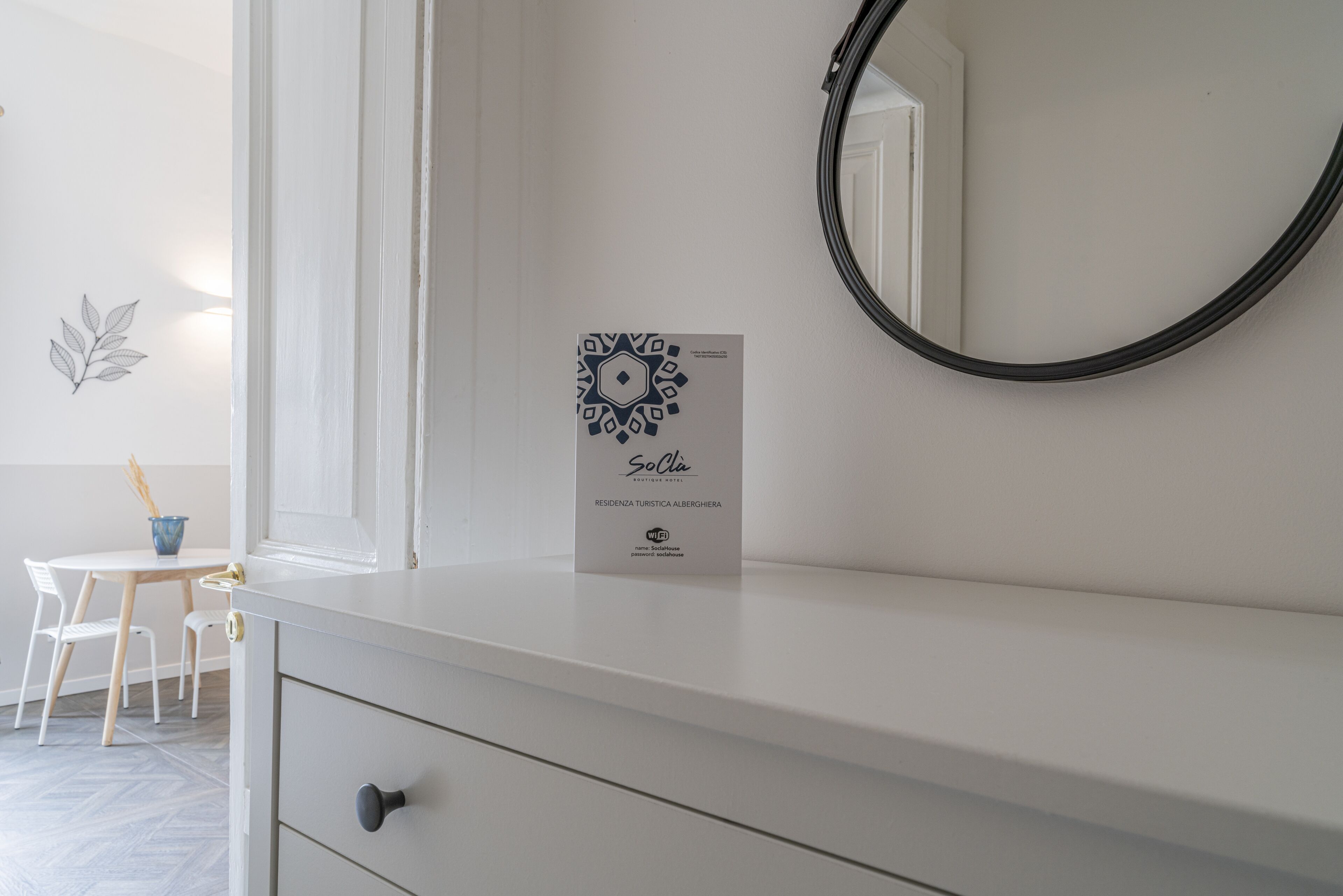 Une commode blanche ornée d’un panneau « SoLia », un miroir rond et une petite table avec des chaises, dans une pièce aux murs blancs et au sol parqueté à motifs en chevrons.