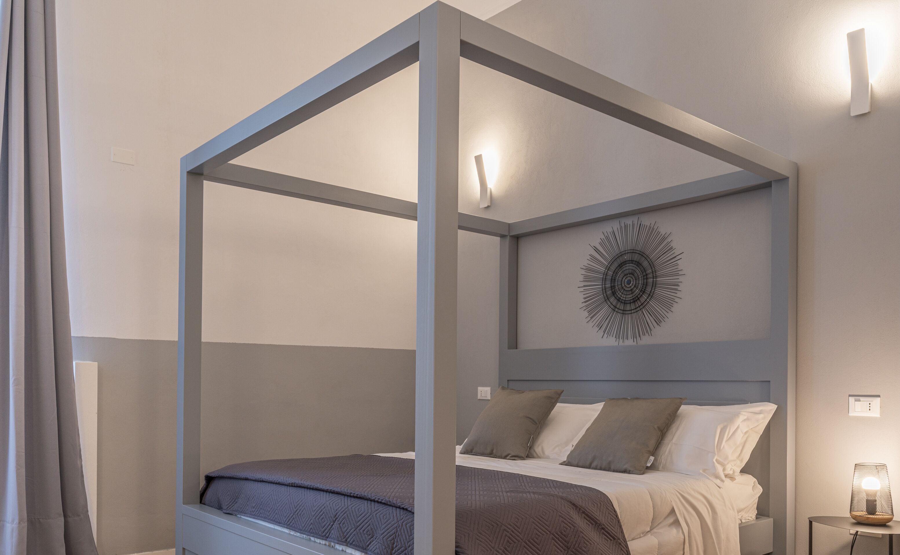A modern bedroom with a four-poster bed, a geometric wall art piece, and a bedside table with a lamp.