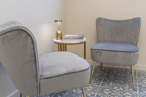 A room with a grey upholstered armchair, a small round table with a gold lamp, and a book titled "COSTA". - SoClà Boutique Hotel (Taranto)