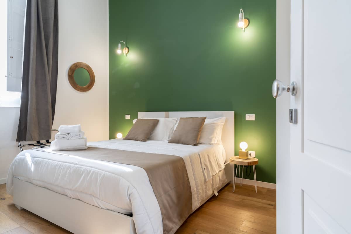Un lit bien fait, recouvert d’une couette blanche et beige, un miroir rond accroché au mur et une table de chevet avec une lampe.