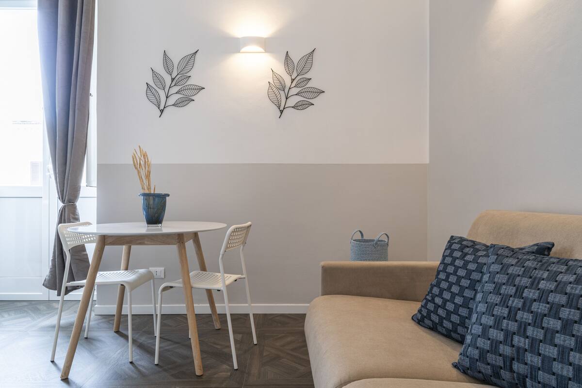 Un salon moderne avec un canapé beige, une petite table ronde entourée de chaises, des œuvres d’art décoratives au mur et une fenêtre avec des rideaux.