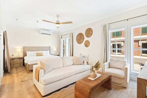 Quarto de hotel com cama, sofá, poltrona, mesa pequena, ventilador de teto e vista para um prédio de tijolos.