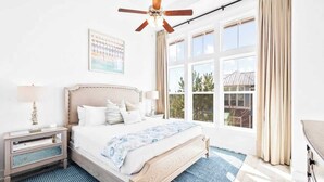 A bedroom with a bed, nightstand, lamp, ceiling fan, large windows, and a view of the outside. - Coastal Oasis by Stay on 30a (Inlet Beach)