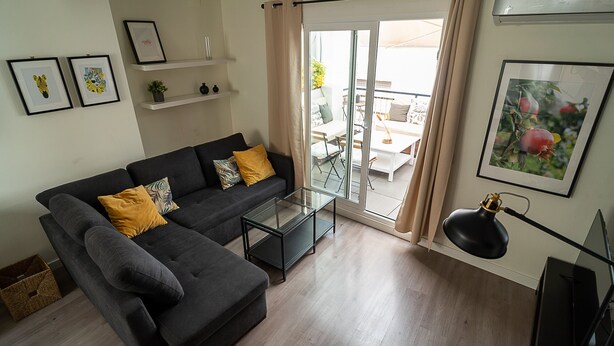 En moderne stue med en grå sofa, et glasbord og udsigt til en udendørs terrasse.