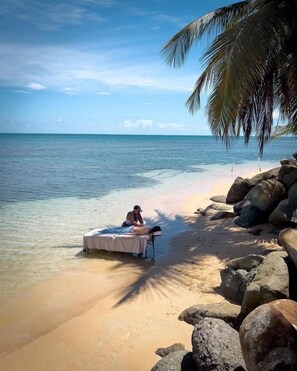 A massage table set up on a beach with clear water and a palm tree in the background.