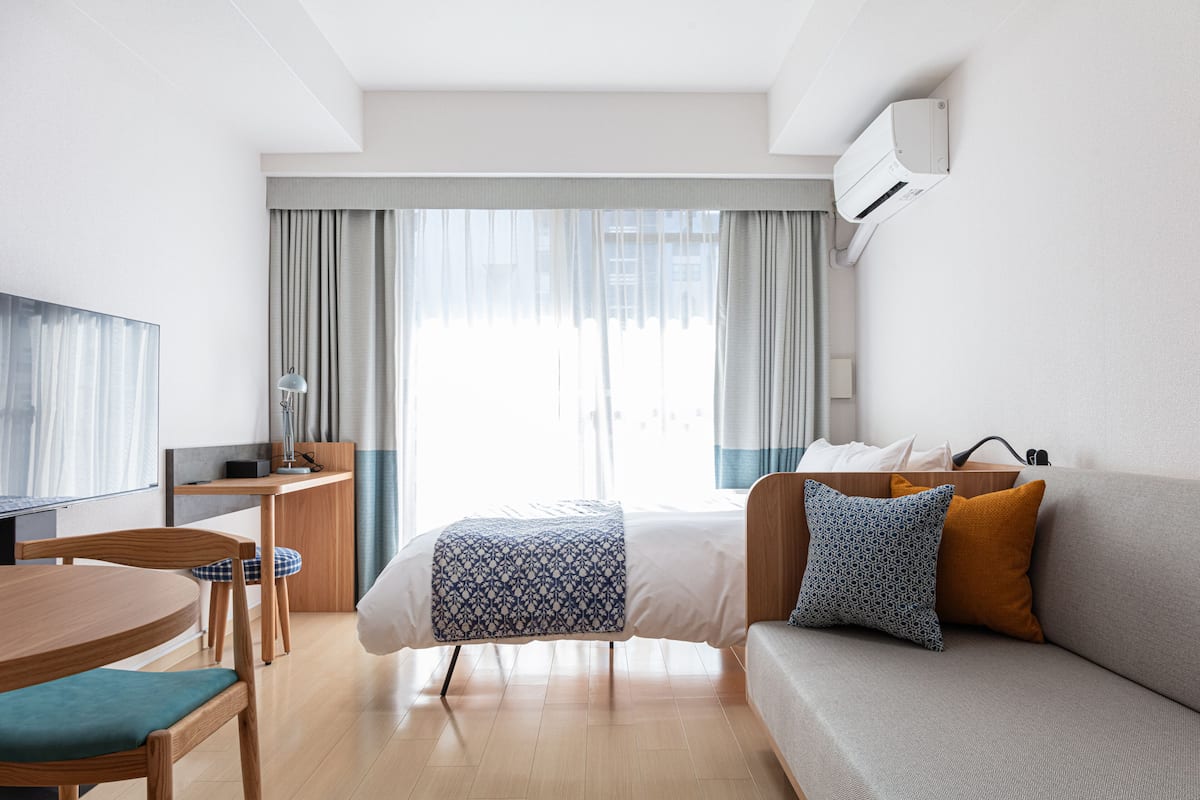 Ein modernes Hotelzimmer mit Bett, Sofa, Schreibtisch mit Stuhl, Fernseher und einem Fenster mit Vorhängen.