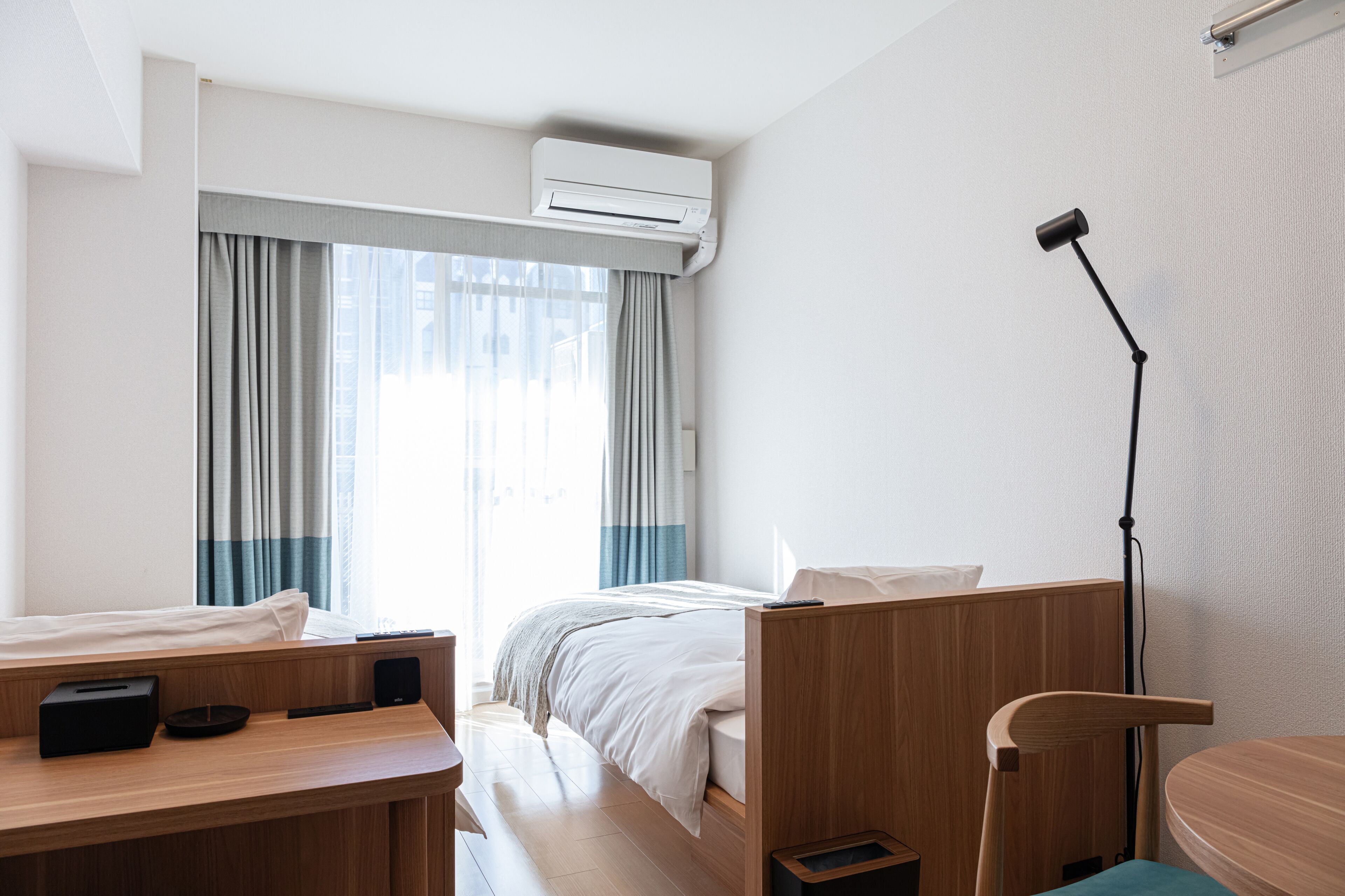 A hotel room with a bed, a desk with a lamp, a chair, and an air conditioning unit.