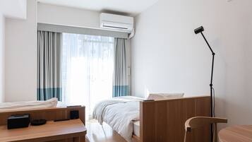A hotel room with a bed, a desk with a lamp, a chair, and an air conditioning unit.