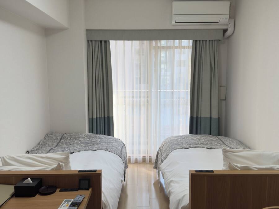 A hotel room with two beds, a desk with a laptop, a lamp, and an air conditioning unit.
