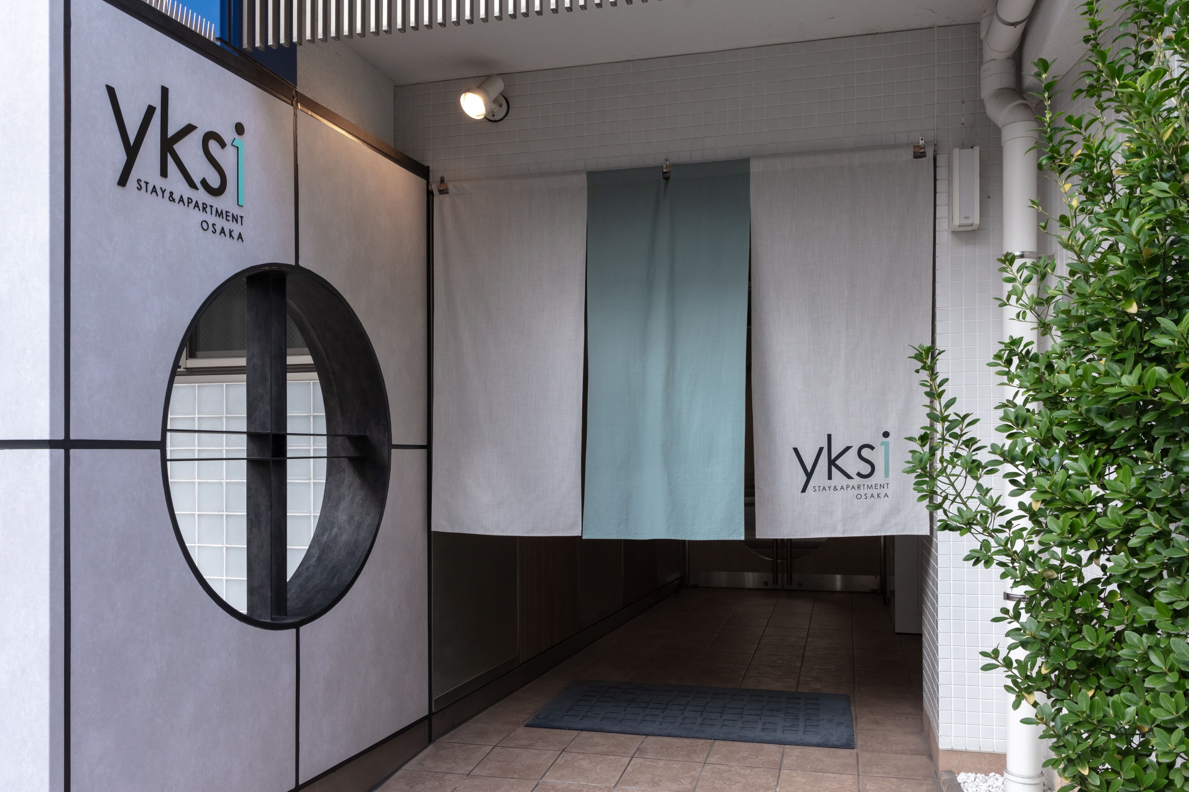 A modern entrance with a circular window, a sign for 'YKSi Stay & Apartment Osaka', and a blue curtain.