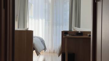 A room with a bed, a desk, and a window with sheer curtains.