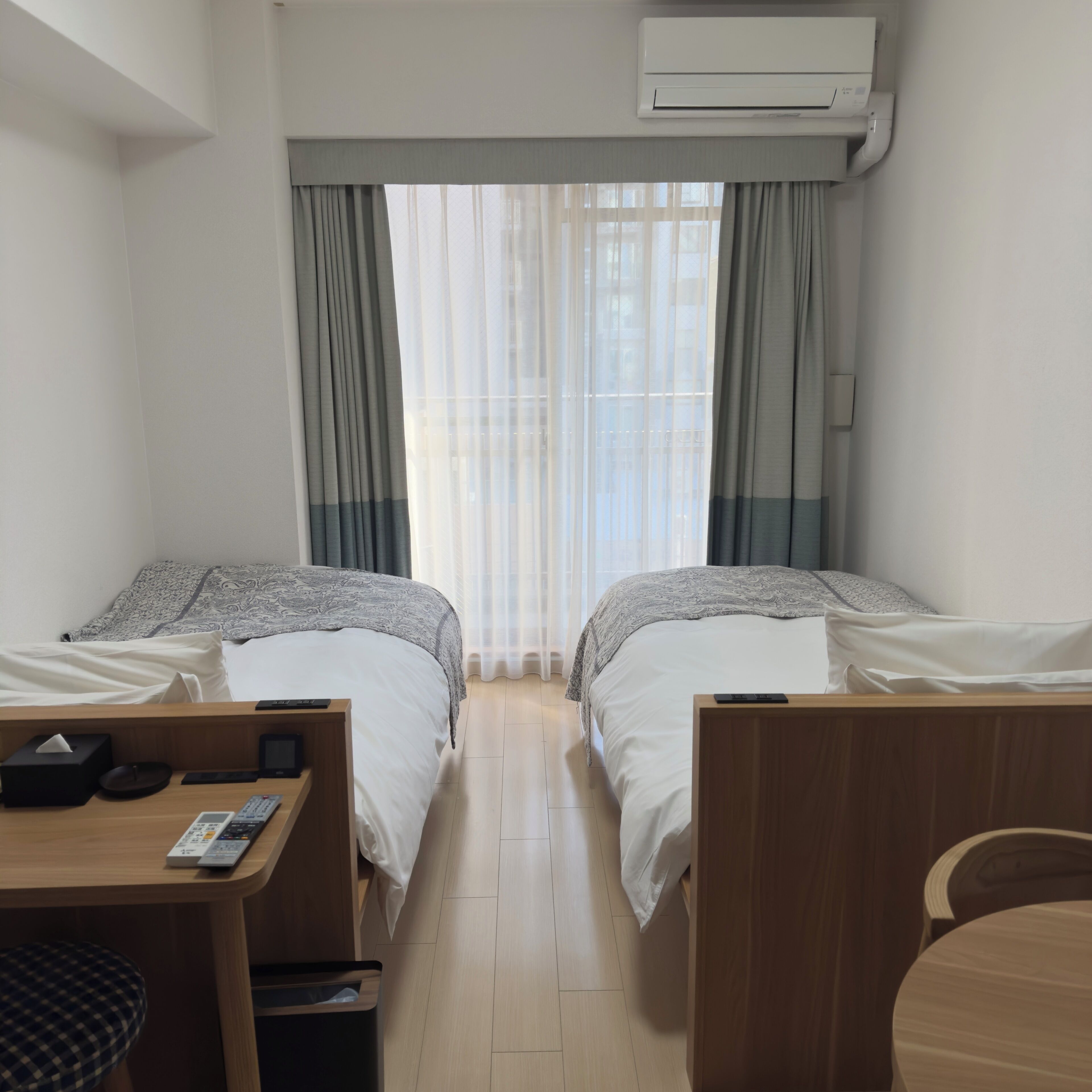 A hotel room with two beds, a desk with a chair, a trash bin, and an air conditioning unit.