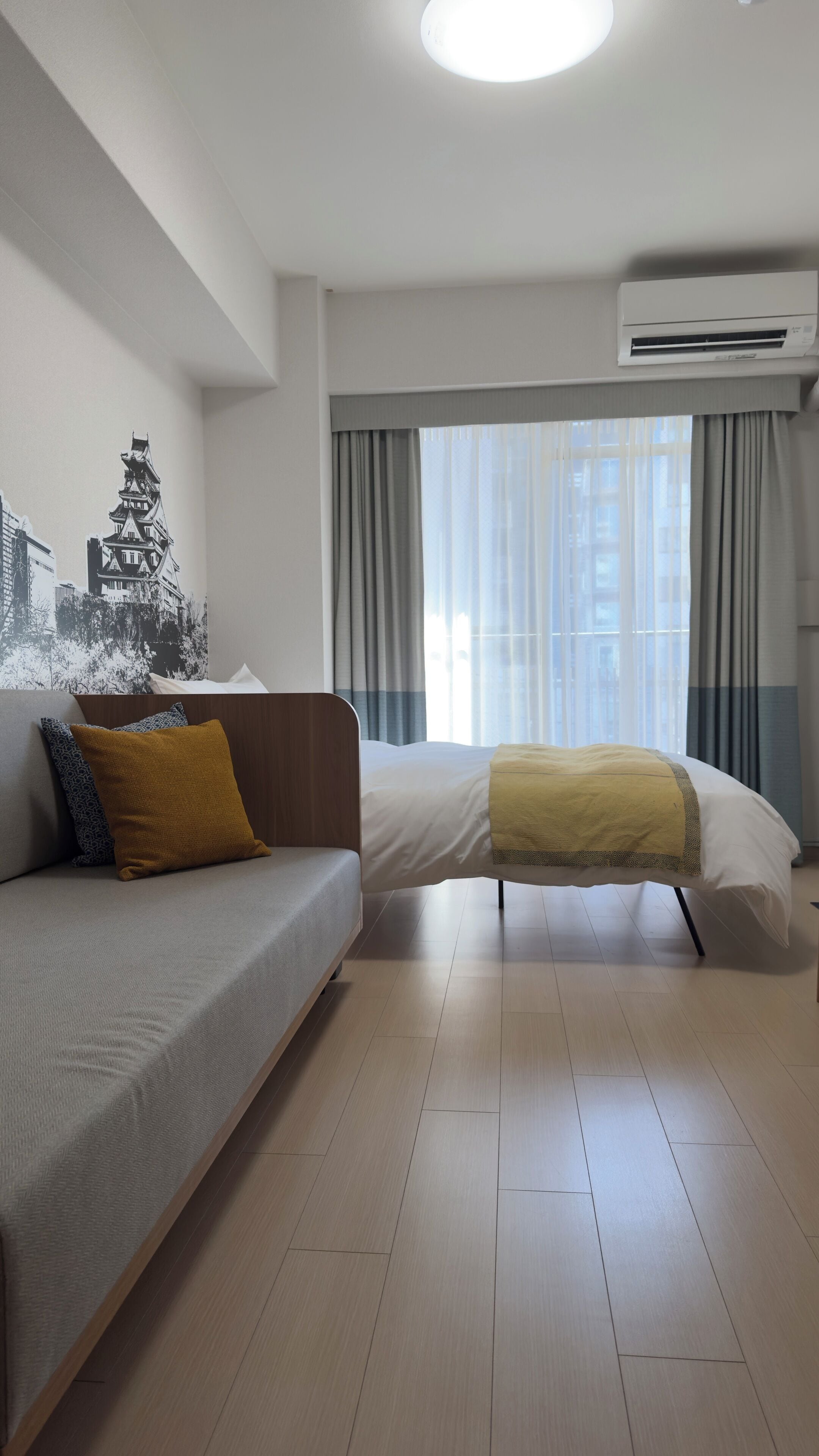 Een moderne hotelkamer met een bank, een bed, een groot raam met gordijnen en een airconditioning.