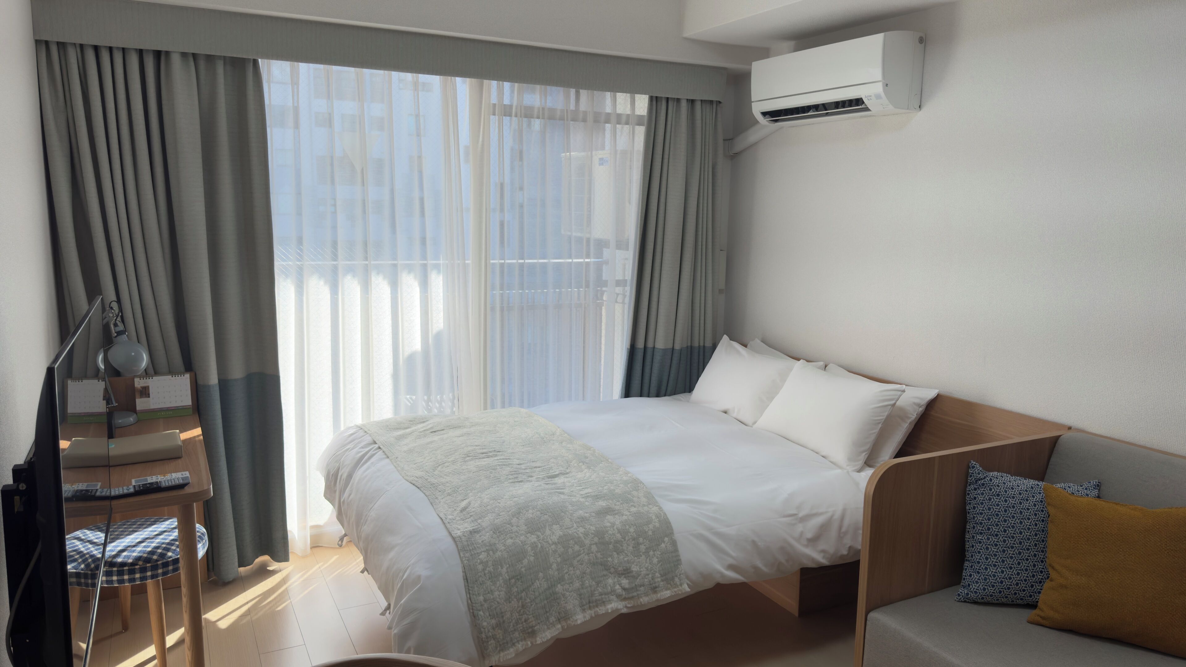 A hotel room with a bed, a desk with a lamp, a chair, and an air conditioning unit.
