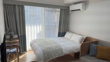 A hotel room with a bed, a desk with a lamp, a chair, and an air conditioning unit.