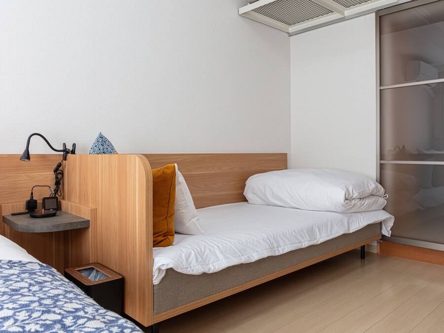 Una habitación de hotel moderna con cabecera de madera, una cama individual con ropa blanca, una mesita de noche con lámpara y un cubo de basura.