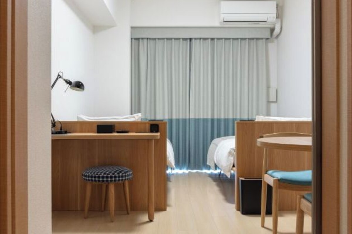 Een compacte hotelkamer met een houten bureau, een klein krukje, een bed met witte beddengoed en een blauw gordijn.