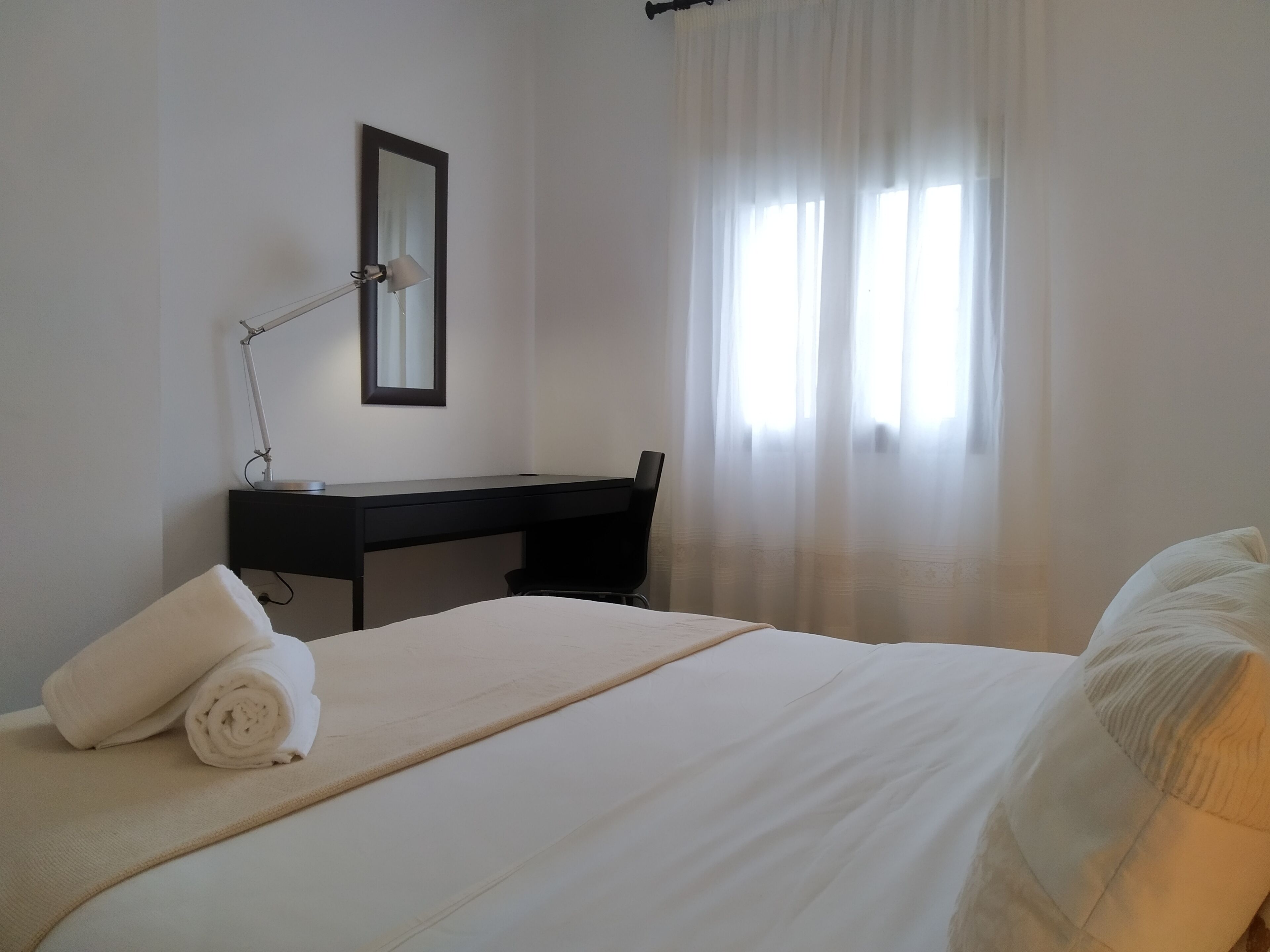A bedroom with a bed, a desk, a chair, a lamp, and a window with curtains.