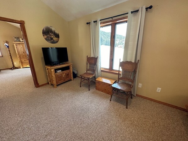 A living room with a television on a wooden stand, two wooden chairs, a wooden chest, and a window with curtains.