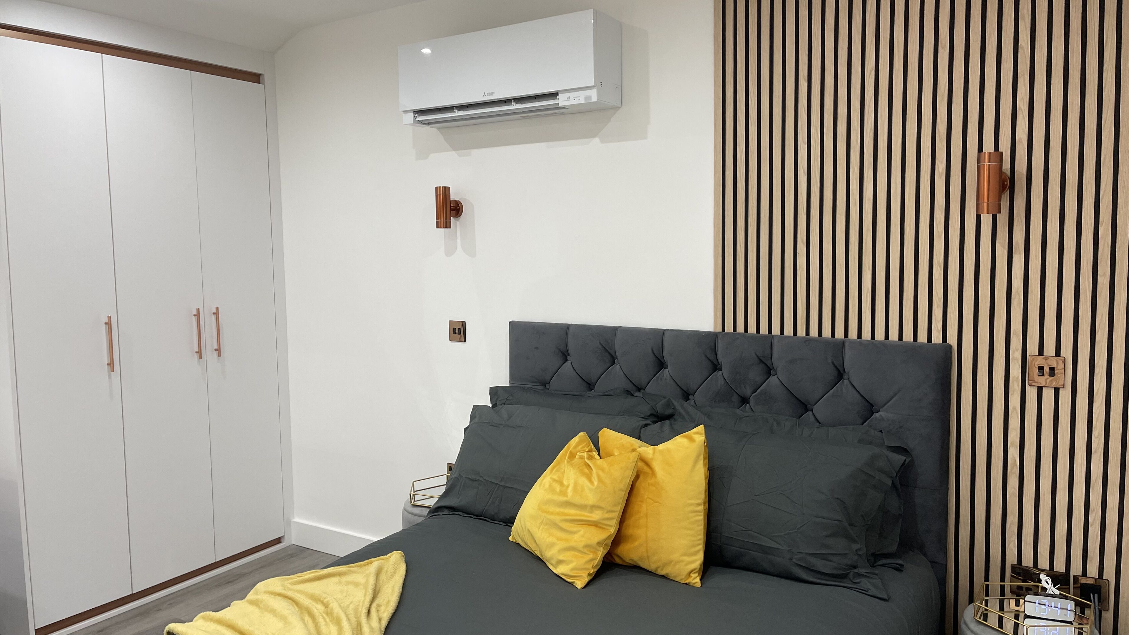 Camera da letto con un letto imbottito grigio, due cuscini gialli, un condizionatore d'aria bianco sopra il letto, un armadio in legno con maniglia marrone e un pannello verticale in legno sulla parete destra.