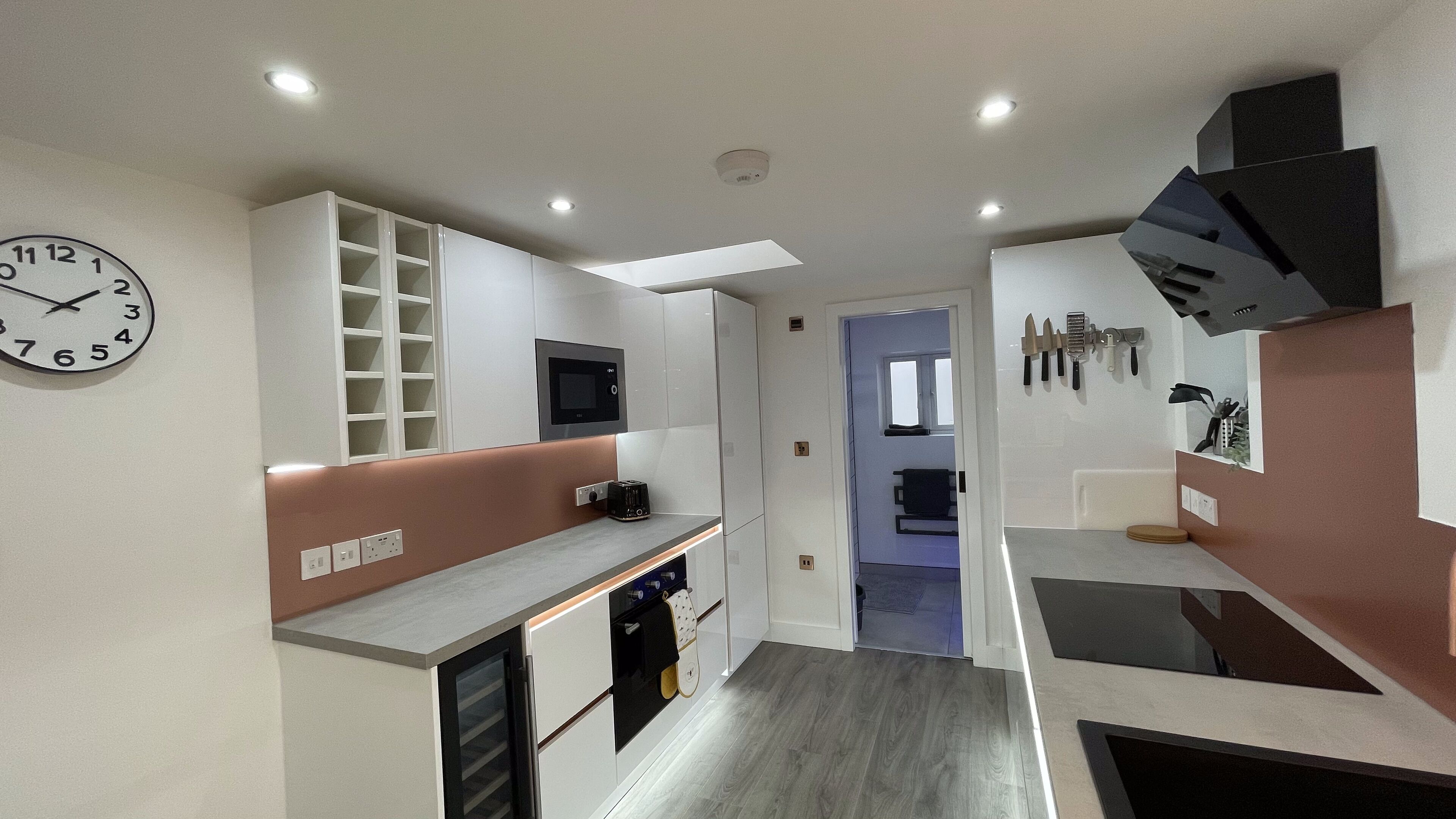 A modern kitchen with white cabinetry, a built-in oven, and a microwave. There is a clock on the wall and a window in the background.