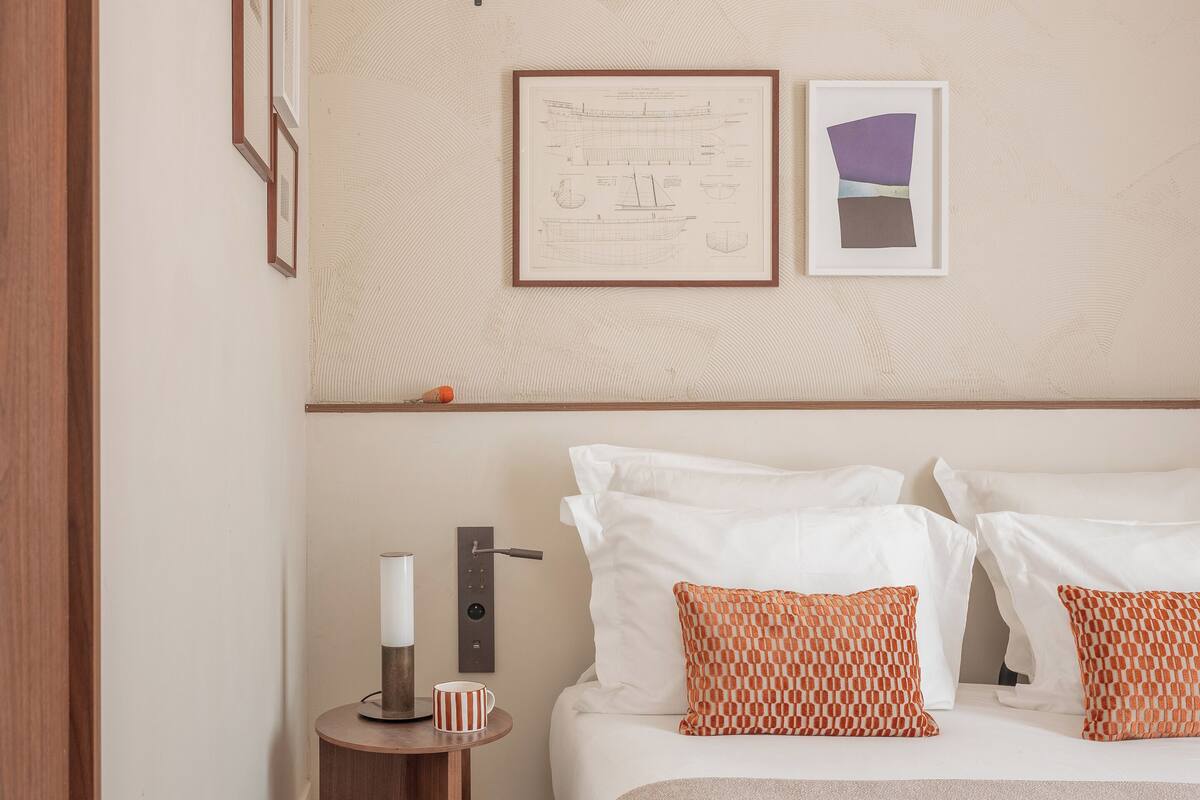 Quarto de hotel com uma cama, almofadas, mesa de cabeceira com abajur, quadro emoldurado e luminária na parede.