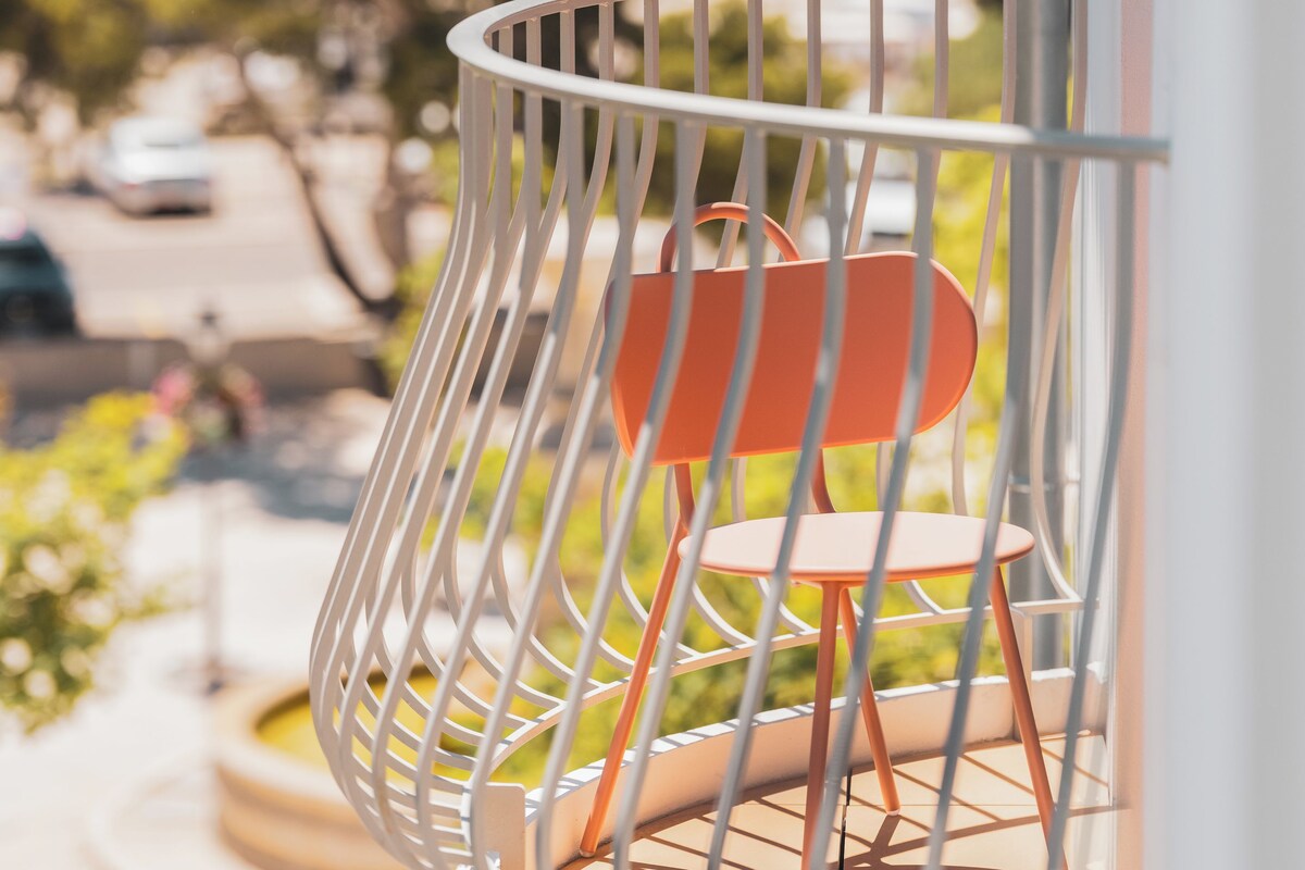 A balcony with a white metal railing, a red chair, and a view of the sea and greenery in the background.