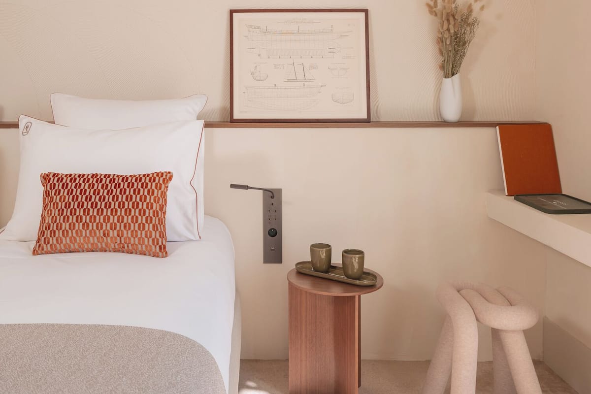 Quarto moderno com uma cama, uma mesinha de cabeceira com duas xícaras, um quadro na parede e um laptop em uma prateleira.