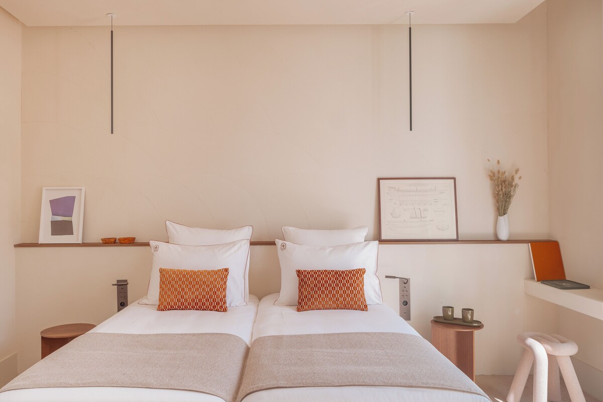 A double bed with white bedding and two orange patterned pillows, a bedside table with a vase of dried flowers, and a framed certificate on the wall.