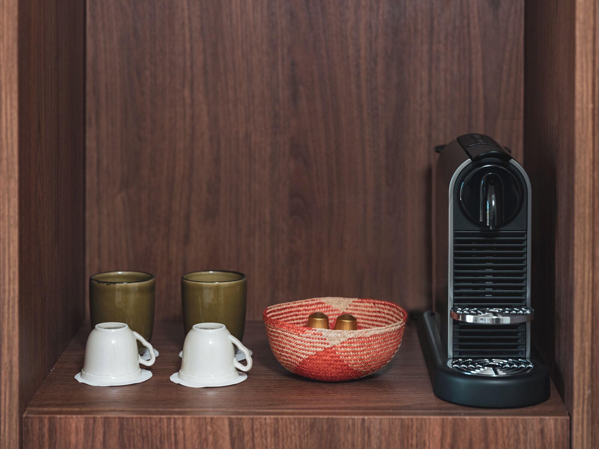Una estantería con una radio negra, una cesta tejida con dos objetos metálicos y dos tazas blancas.
