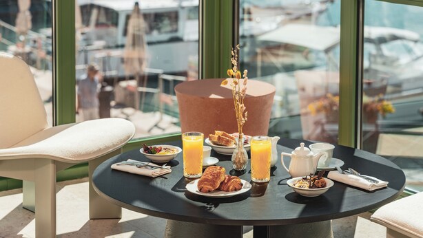 A round table with breakfast items, a vase with flowers, and a view of boats through large windows. - Hôtel Liautaud Cassis by Germain Collection (Cassis)