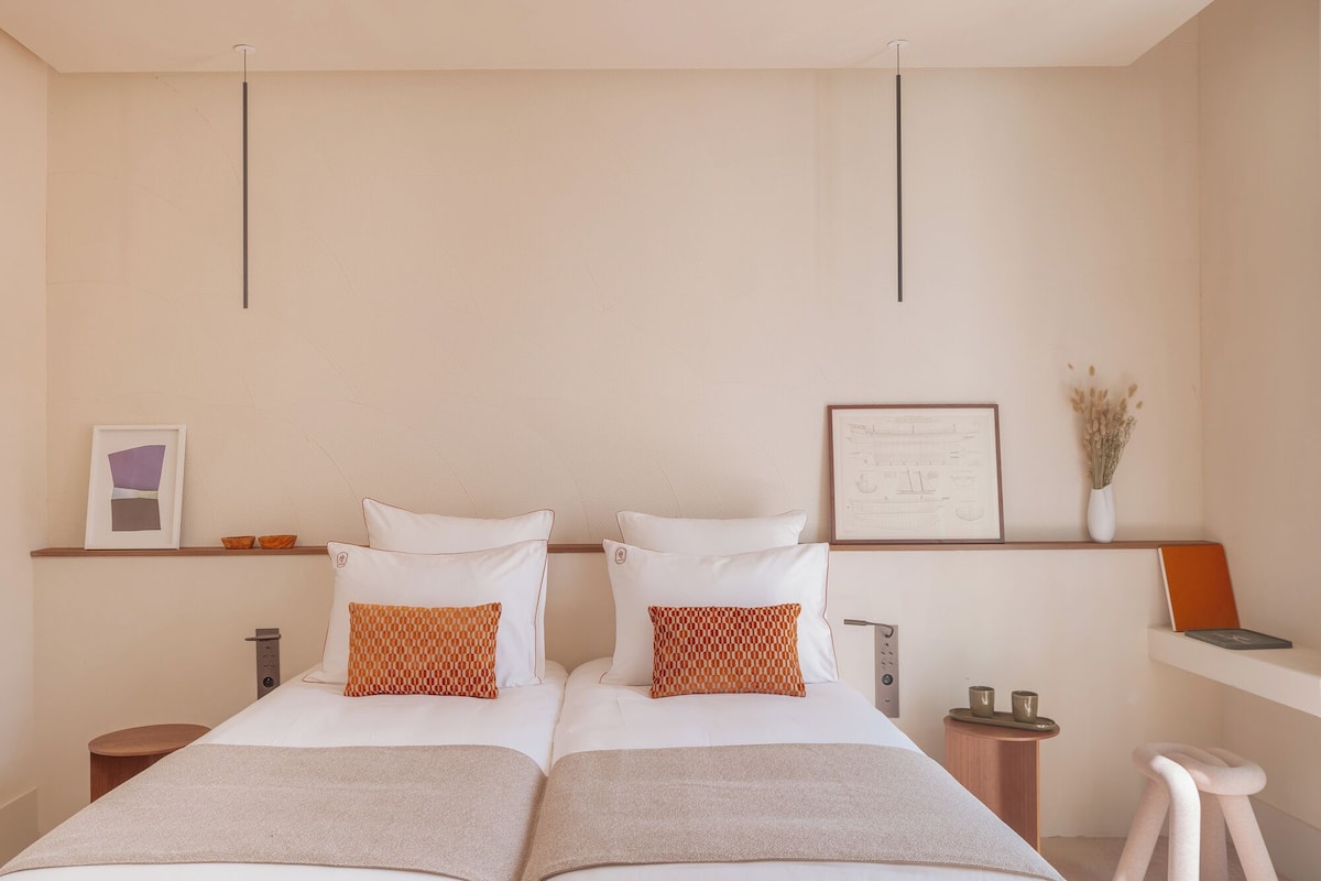 Quarto de hotel com duas camas, uma mesa de cabeceira de madeira, um certificado emoldurado na parede e um banquinho pequeno.