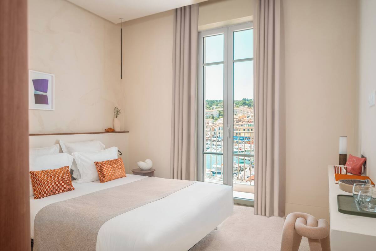 Quarto de hotel com uma cama grande, uma mesa com cadeira e vista para a paisagem urbana através de uma janela ampla.