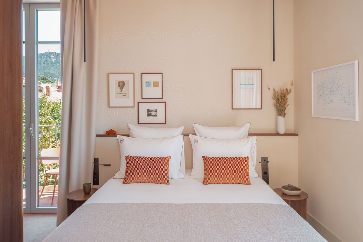 A hotel room with a large bed, two pillows, a nightstand, a vase with dried flowers, and framed pictures on the wall.