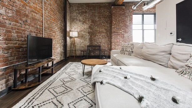 A room with a brick wall, a flat-screen TV on a wooden stand, a round wooden table, a white sofa, and a patterned rug. - Loft in the Rivermarket District! Rooftop access and DT KC views. Walk to City Market, local coffee shops, restaurants, shops or hop on the free streetcar just steps away. Minutes from  P&L, T-Mobile, Crossroads, Plaza. 10 min drive to stadiums. (Kansas City)