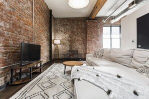 Une pièce avec un mur en briques, un téléviseur à écran plat posé sur un meuble en bois, une table ronde en bois, un canapé blanc et un tapis à motifs.