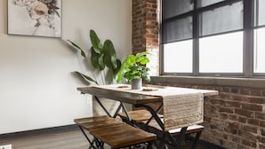 A dining area with a wooden table, woven chair backs, and a potted plant. - Exposed brick, wood beams, & chic touches give this loft serious style. Enjoy skyline views from the rooftop, a full kitchen with coffee bar, king bed, on-site gym, & quick access to the free KC streetcar. Walk to shops/dining, and the City Market! (Kansas City)