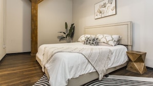 A bedroom with a wooden ceiling, a bed with a white headboard, a framed floral artwork, and a geometric side table. - Exposed brick, wood beams, & chic touches give this loft serious style. Enjoy skyline views from the rooftop, a full kitchen with coffee bar, king bed, on-site gym, & quick access to the free KC streetcar. Walk to shops/dining, and the City Market! (Kansas City)