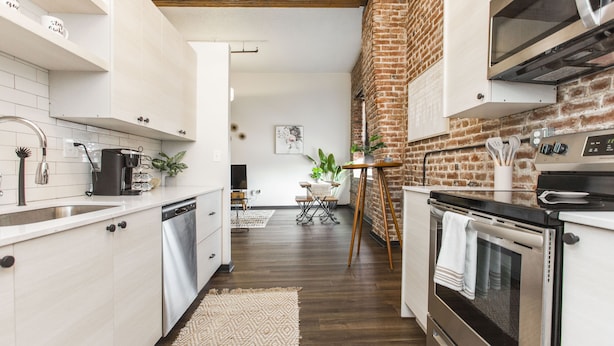 A modern kitchen with white cabinetry, a brick accent wall, stainless steel appliances, and a dining area in the background. - Exposed brick, wood beams, & chic touches give this loft serious style. Enjoy skyline views from the rooftop, a full kitchen with coffee bar, king bed, on-site gym, & quick access to the free KC streetcar. Walk to shops/dining, and the City Market! (Kansas City)