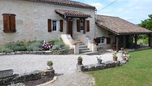 Une maison en pierre avec un toit en tuiles, un porche couvert et une allée de gravier agrémentée de plantes en pot.