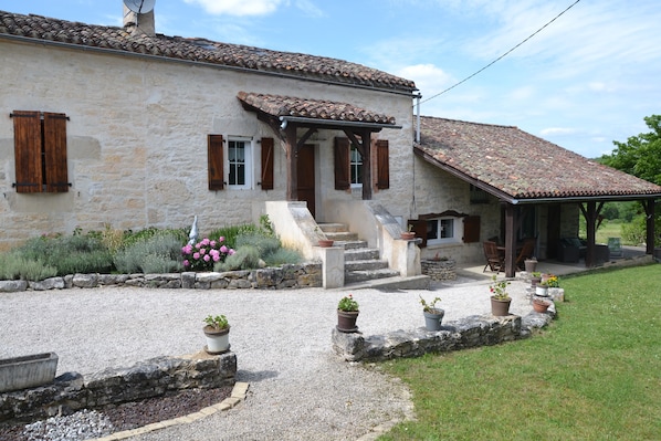 A stone house with a tiled roof, a covered porch, and a gravel driveway with potted plants.