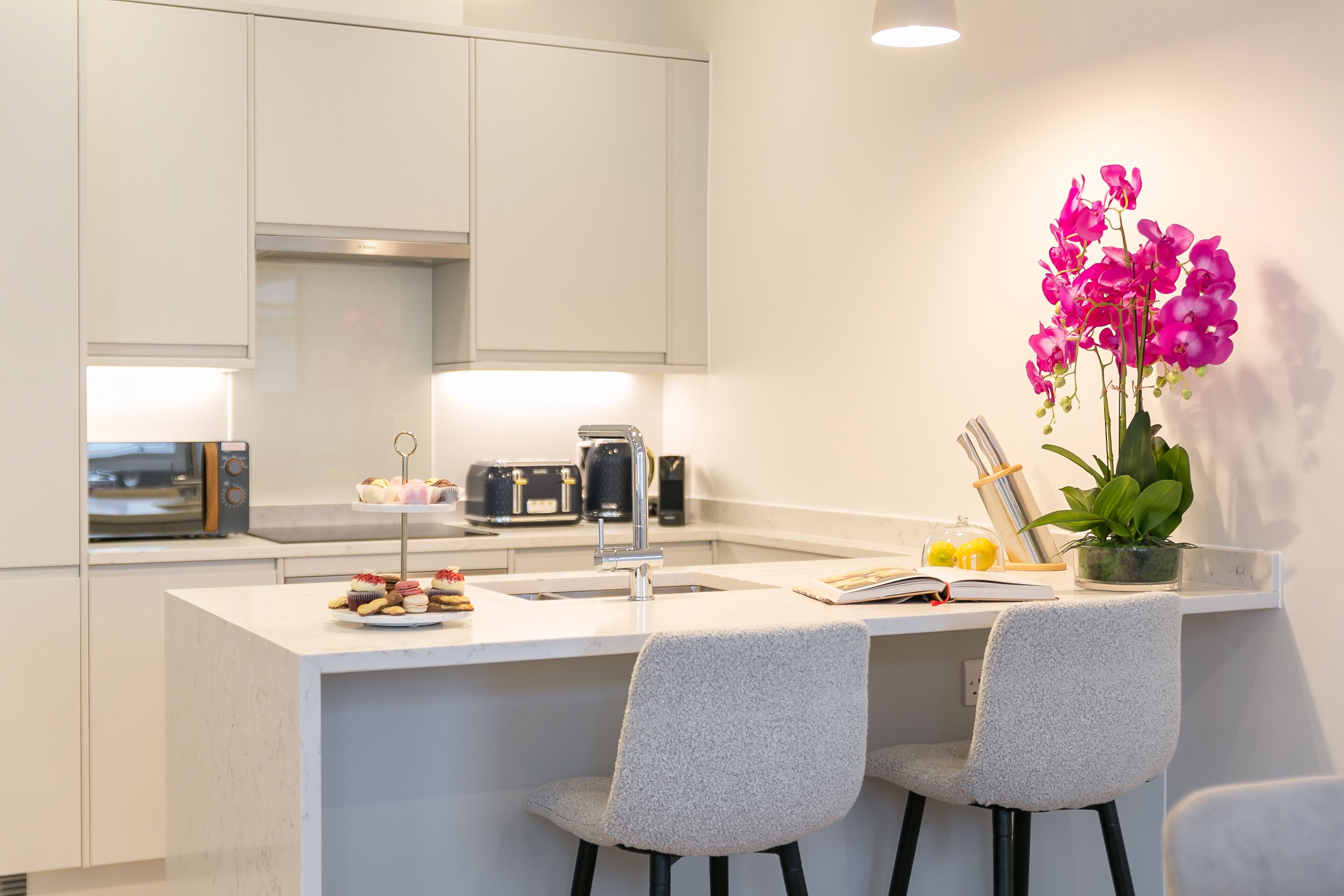 Une cuisine moderne avec un îlot central, deux chaises grises, un évier et une orchidée en pot.