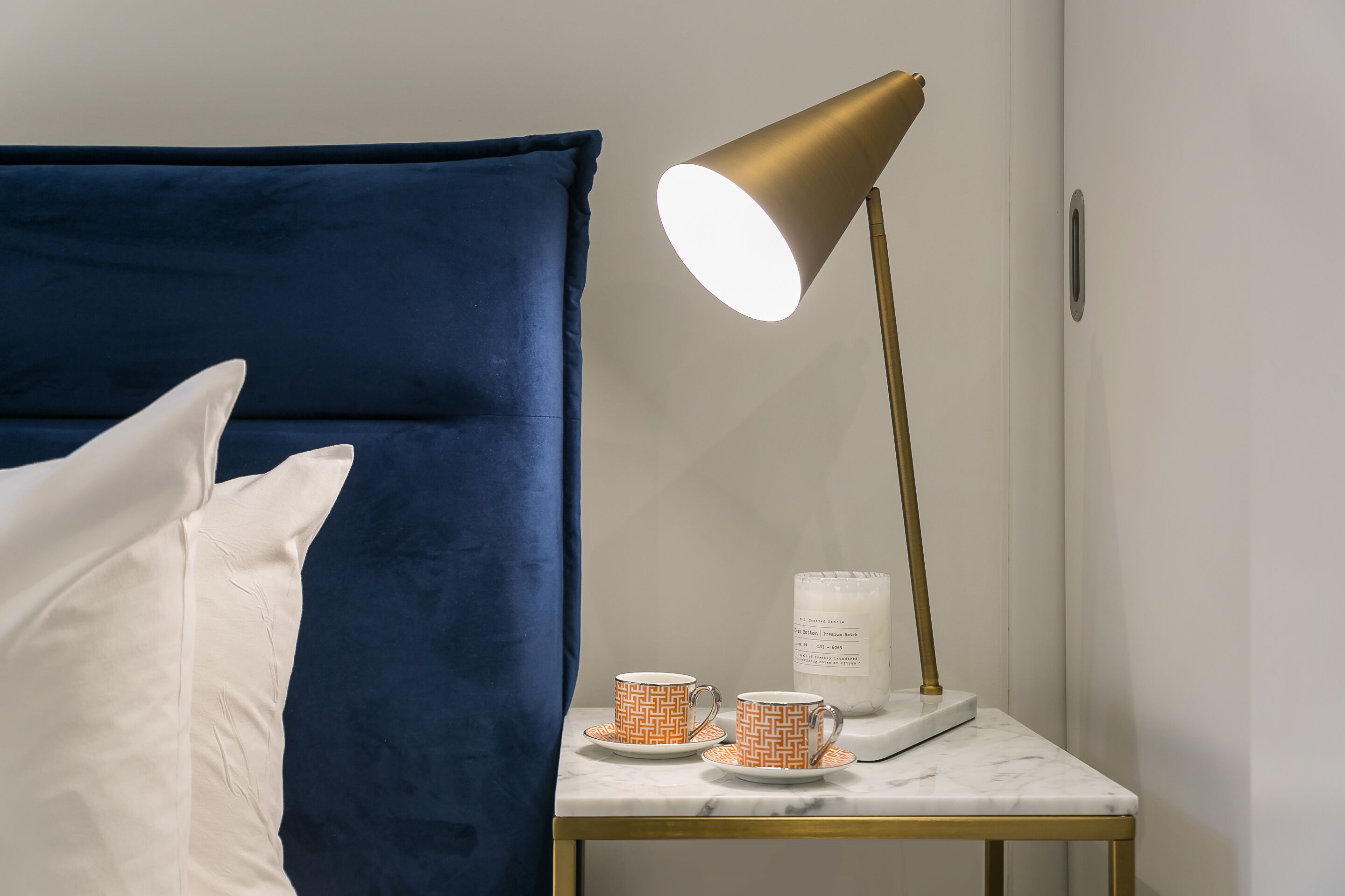 Un lit avec des oreillers blancs, une tête de lit bleue, une table de chevet avec une lampe dorée et deux tasses à motifs posées sur la table.