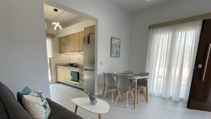 A modern living room with a sofa, a dining table with chairs, a kitchen in the background, and a large window with curtains.