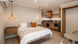 A modern bedroom with a large bed, bedside tables, a small table, and a chair. There is a kitchen area with cabinets and appliances.