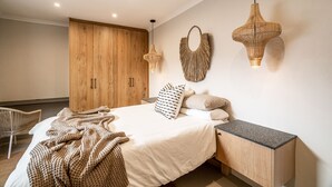 A bedroom with a bed, wooden wardrobe, woven wall decor, and a bedside table. - View 16 | Serinity Skyline Penthouse | 3 Bedroom (Cape Town)