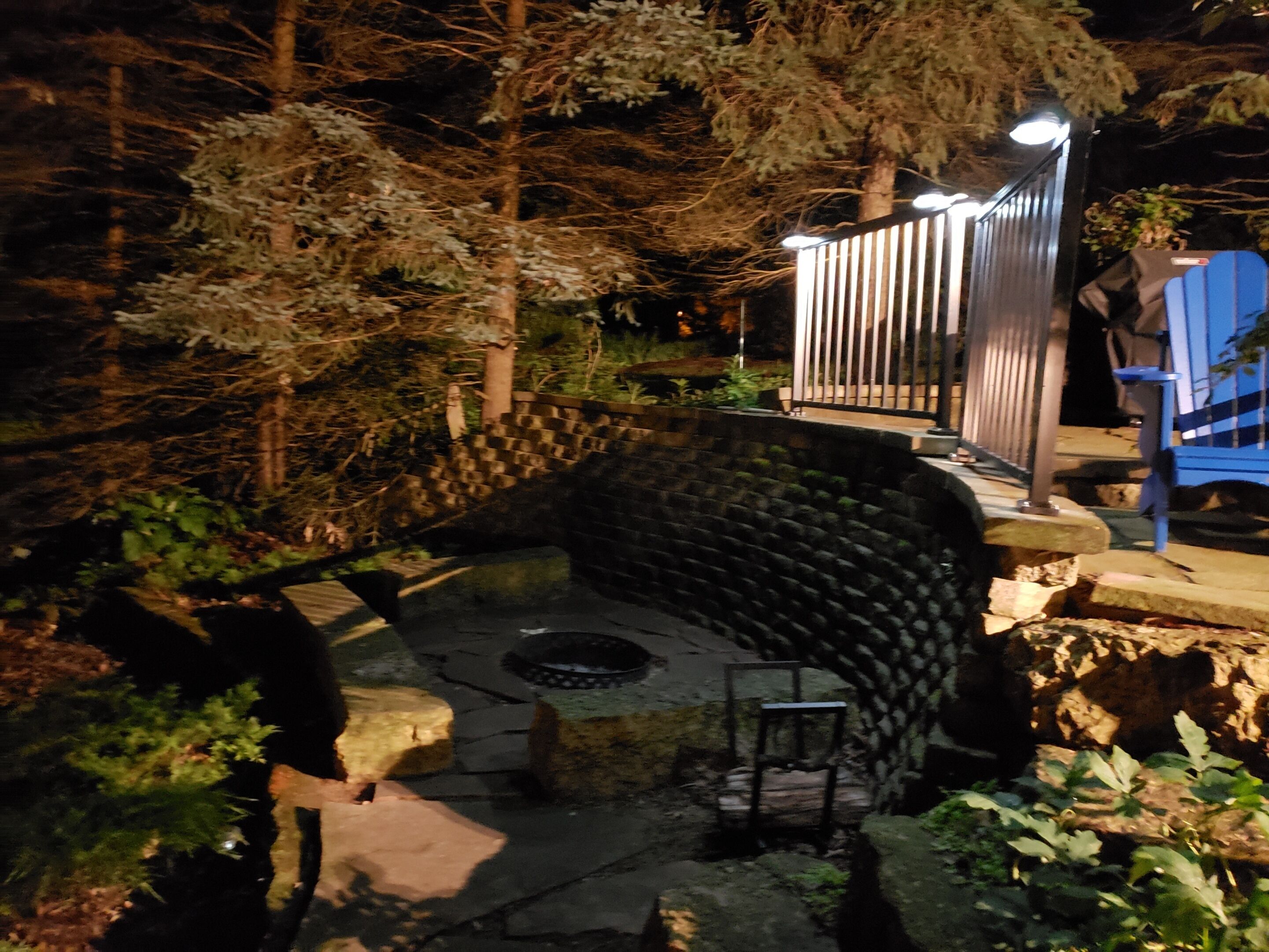 Un espace extérieur aménagé avec des sièges en pierre, doté d’un foyer, et entouré d’arbres et d’une clôture en métal.